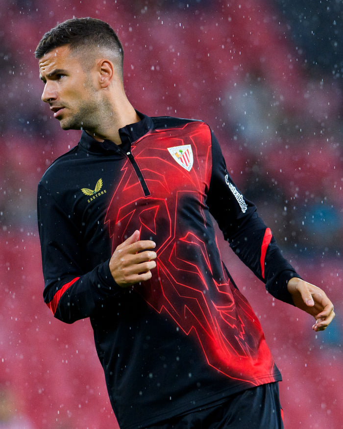 Athlete wearing MATCHDAY MIDLAYER 24/25 jacket in black with red geometric pattern and club crest in the rain.