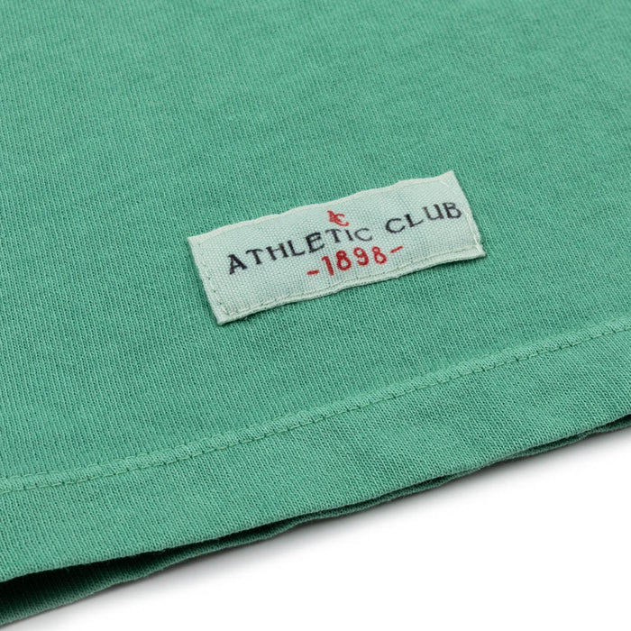 Close up of green Junior AC Flag Tee fabric with a white label reading Athletic Club 1988