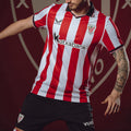 Model wearing Athletic Club home shirt with bold red and white stripes and crest on chest