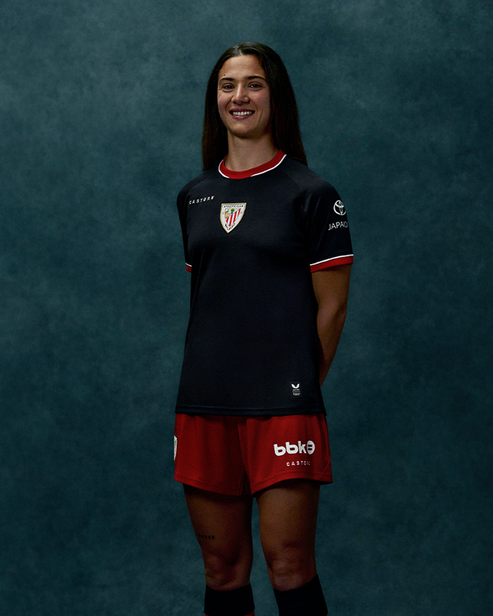 Front view of a woman wearing a black away short sleeve soccer jersey with red trim and club crest on the chest.