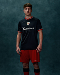 Male model wearing black away pro short sleeve soccer jersey with red shorts and Kutxabank sponsor against teal backdrop