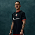 Male model wearing black away short sleeve shirt with red trim posing against teal backdrop