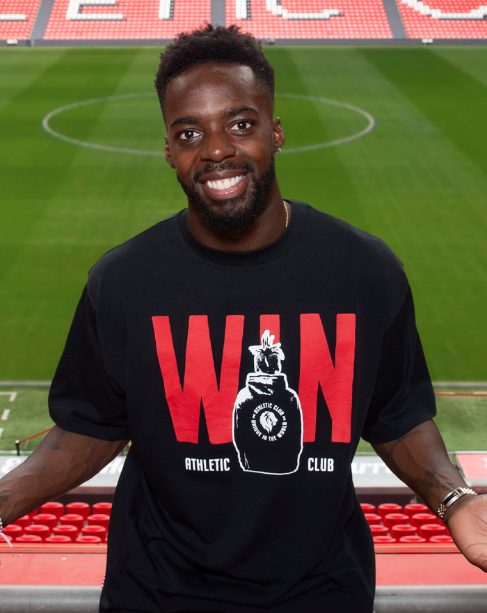 Smiling man models a black Nico Win graphic T shirt at a stadium.