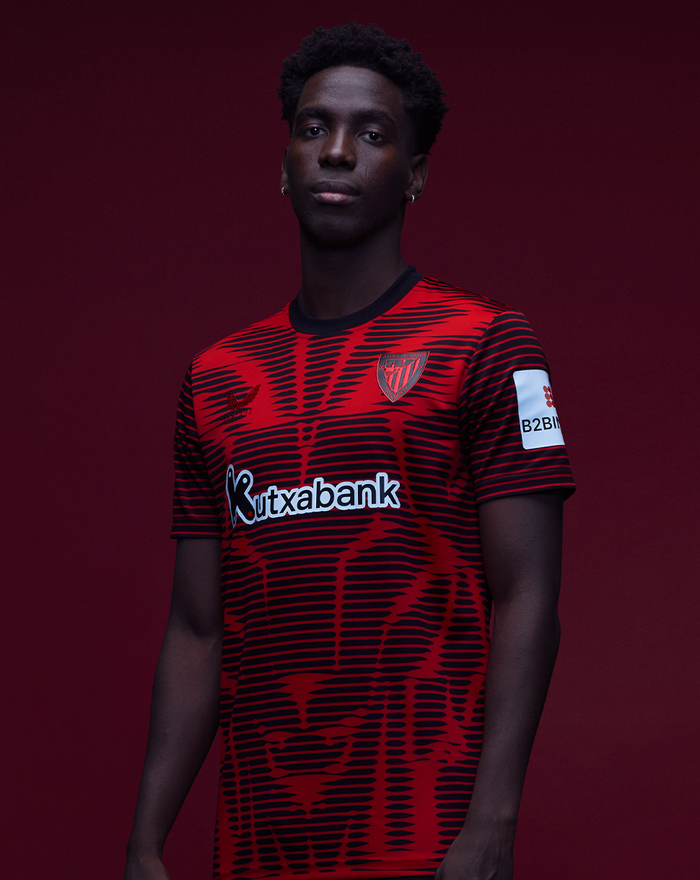 Male model wearing Athletic Club 4th short sleeve shirt with red and black pattern against a dark red studio backdrop.
