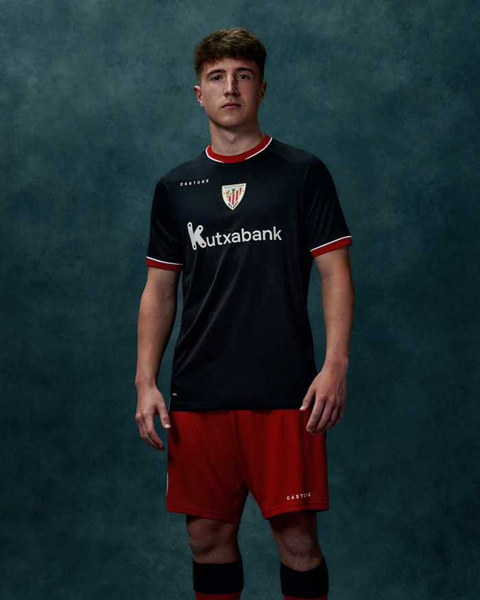 Male model wearing black away short sleeve shirt with red trim and red shorts against a teal textured backdrop